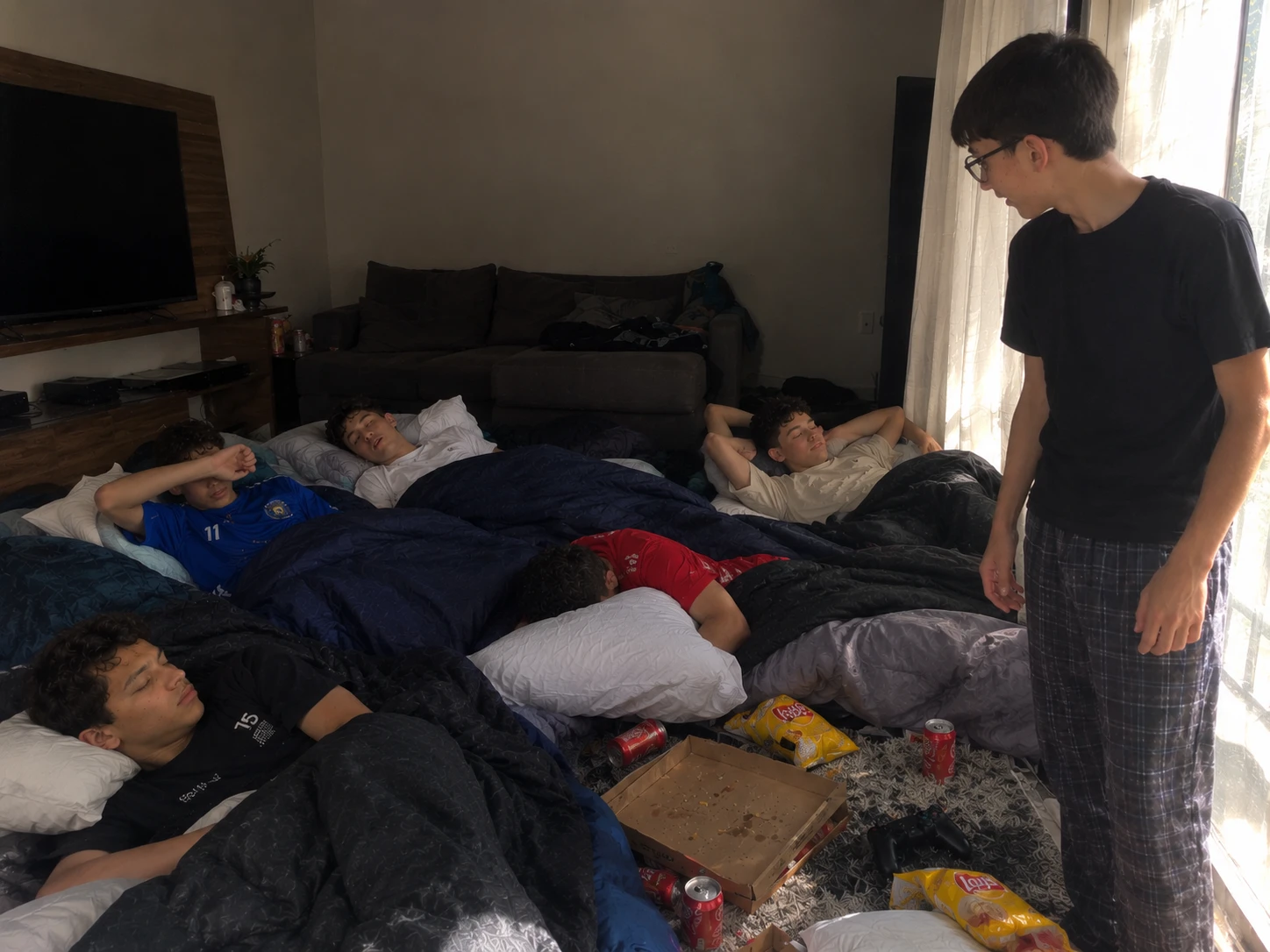 Foto de verdade: de manhã, numa sala com TV e sofá, cinco amigos dormem em colchões no chão, cercados de caixa de pizza vazia, latinhas de refrigerante, salgadinho e um controle de videogame. Bertini, de óculos e pijama, está de pé olhando para eles.